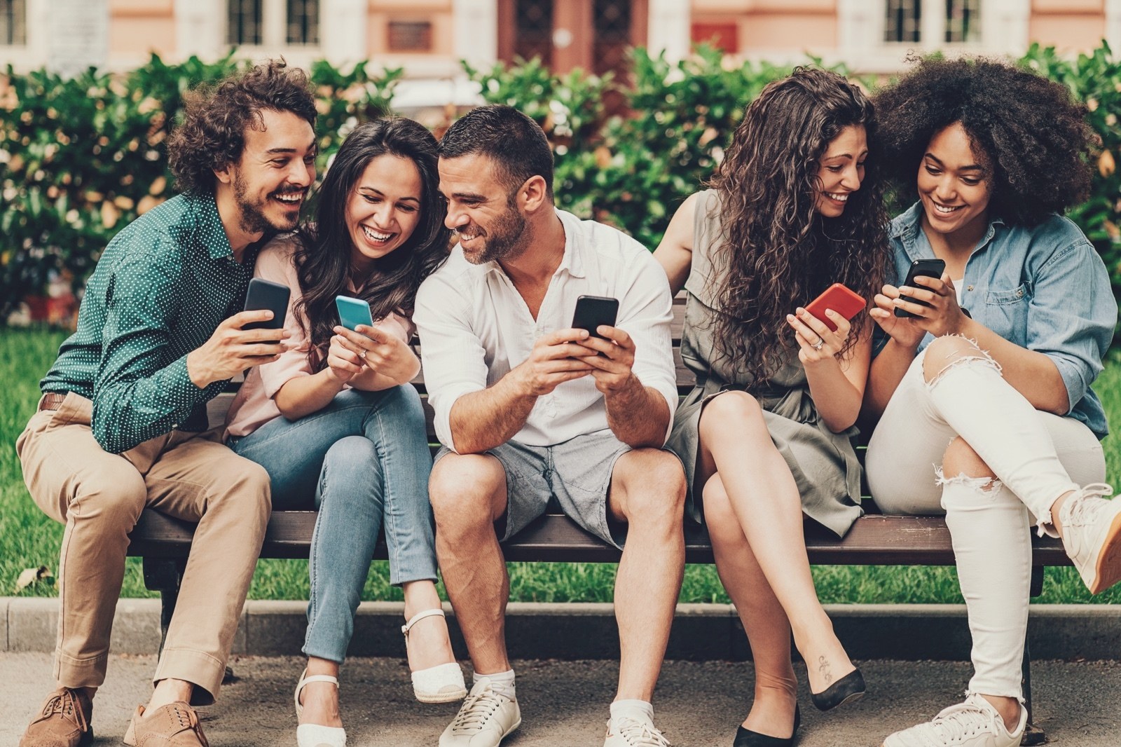 Cinco jovens sentados num banco de praça, rindo enquanto olham o celular