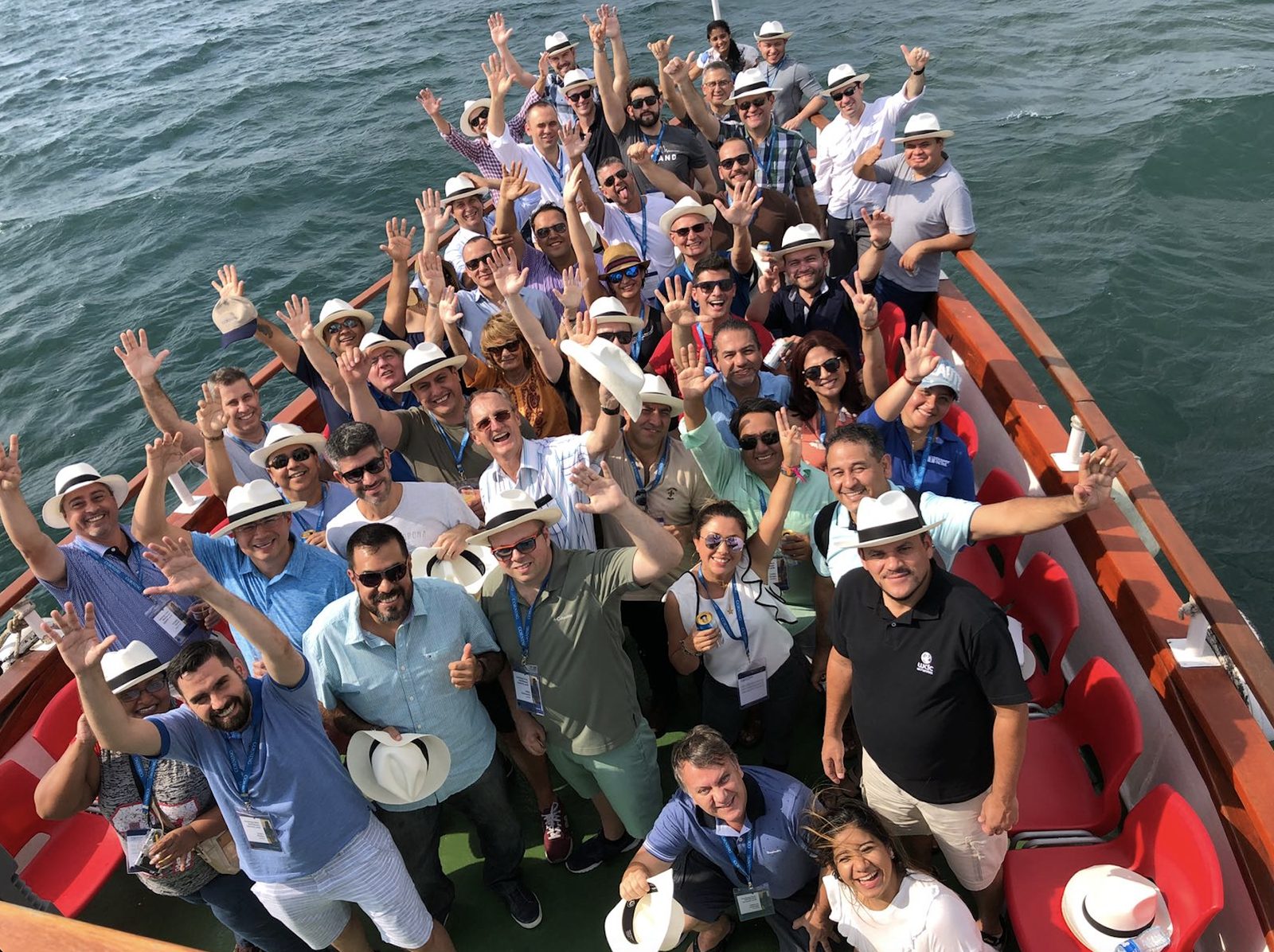 Participantes do Corning CALA Channel Summit reunidos na proa de um barco, de chapéu panamá, acenando para a câmera